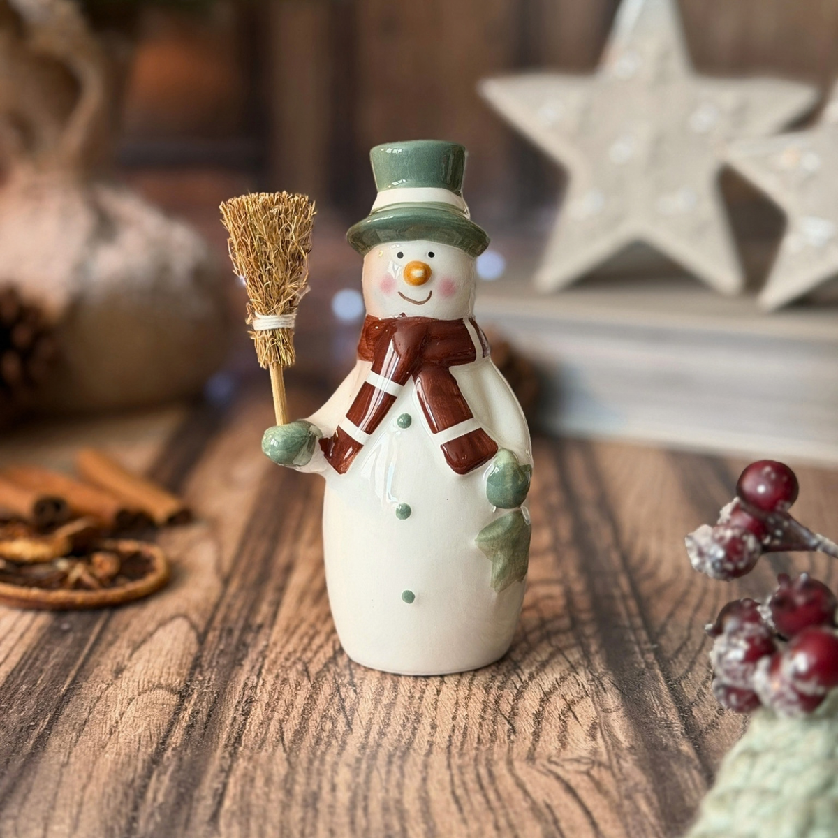 A ceramic snowman figurine holding a small broom, decorated with a green top hat, brown scarf, and green mittens, displayed on a wooden surface with holiday-themed background props.