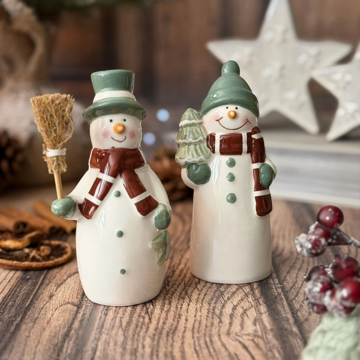 Two ceramic snowman on a rustic wooden table, decorated with green hats, brown scarves, and holding a small broom and a miniature Christmas tree.