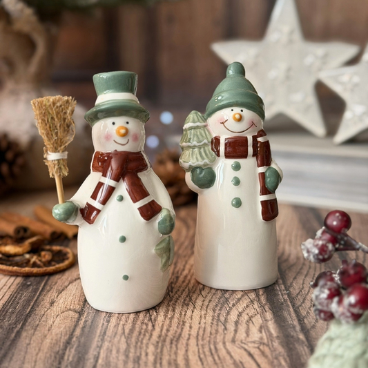 Two ceramic snowman on a rustic wooden table, decorated with green hats, brown scarves, and holding a small broom and a miniature Christmas tree.