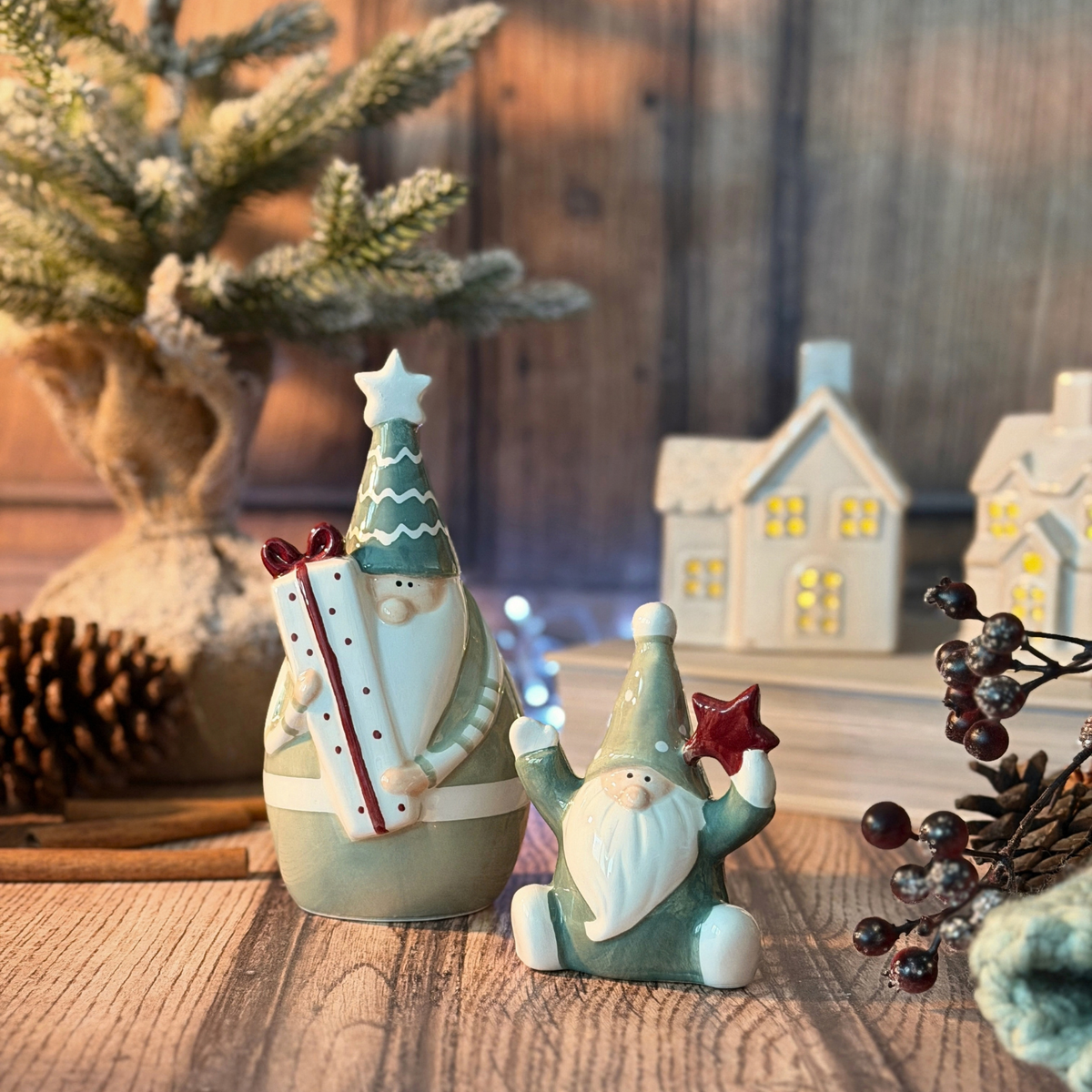 Two ceramic Christmas santas on a wooden surface, with a blurred rustic holiday scene in the background including small house lights, pinecones, and frosted greenery.