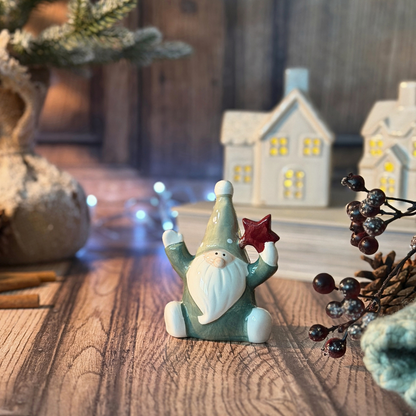 A small ceramic santa figurine with a green body, white beard, and red star, displayed on a wooden surface with cozy holiday decorations in the background.