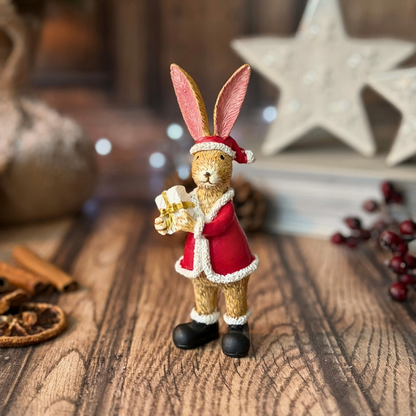 A small decorative rabbit figurine dressed in a red Santa-style coat and hat, holding a wrapped gift, displayed on a wooden surface with holiday props in the background.