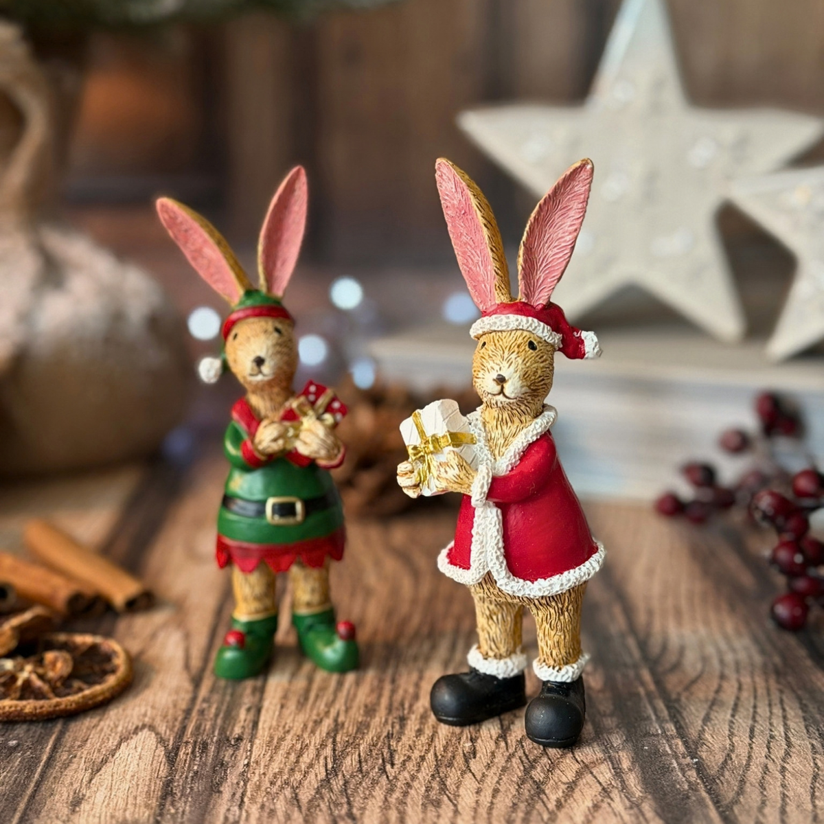 Two Christmas-themed rabbit figurines standing on a wooden surface. One rabbit wears a green elf outfit with red trim and holds a wrapped gift, while the other wears a red Santa-style coat with white trim and holds a small box.