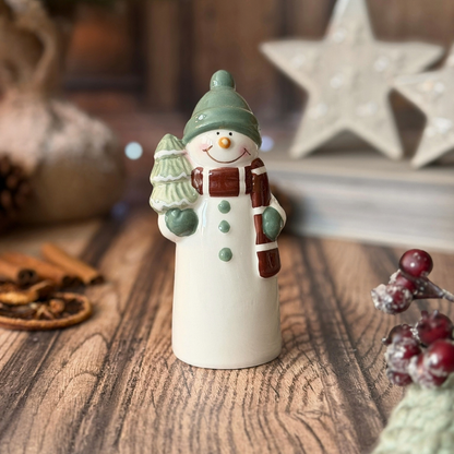 A ceramic snowman figurine standing on a wooden surface, wearing a green hat, green scarf details, and a brown-and-white striped scarf, with a small green tree on one hand. The background is softly blurred with holiday decor, including a star and berries.