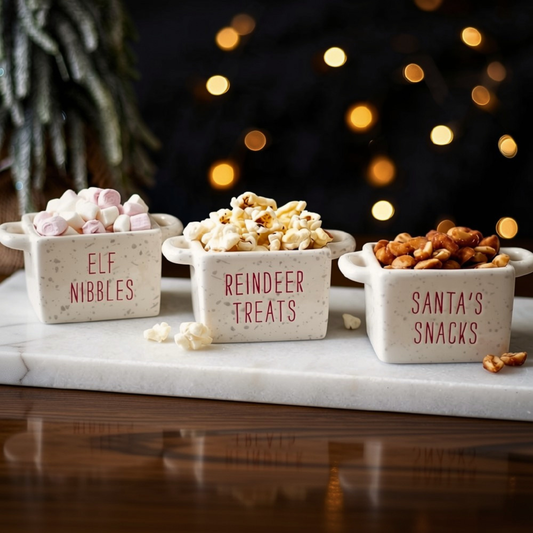 Three small speckled cream ceramic snack bowls labeled 'ELF NIBBLES,' 'REINDEER TREATS,' and 'SANTA'S SNACKS' filled with marshmallows, popcorn, and peanuts, arranged on a white marble board with a dark festive background and warm string lights.