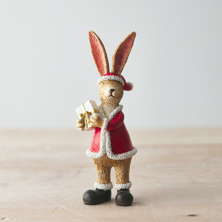 Decorative rabbit figurine in a Santa outfit holding a gift on a wooden surface.
