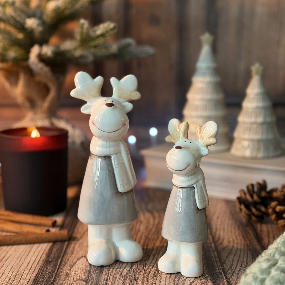 Two ceramic Christmas reindeer figurines in light gray and white, standing on a rustic wooden surface with a lit candle, pinecones, and blurred holiday decorations in the background.