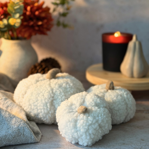 Three small white faux-fur decorative pumpkins arranged on a tabletop with a linen napkin, a vase of flowers, pinecones, and a lit black candle in the background.