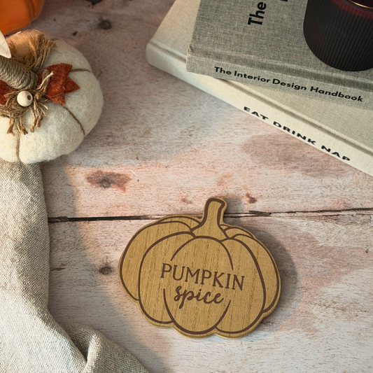 A wooden pumpkin-shaped sign engraved with 'Pumpkin Spice' sits on a rustic whitewashed wooden surface, styled with a linen cloth, a small fabric pumpkin, and books in the background.