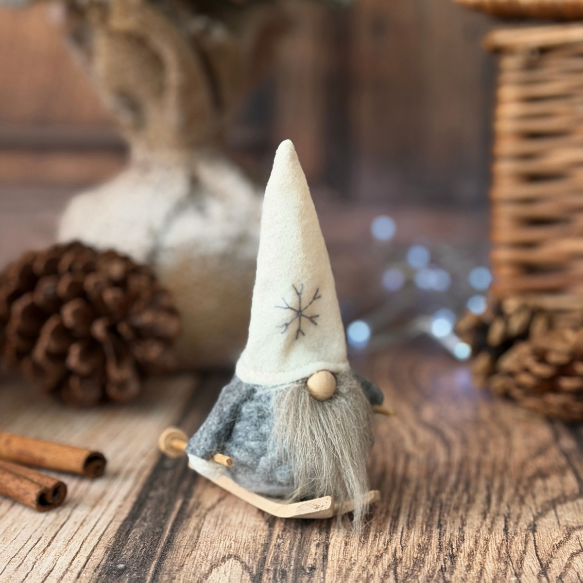 A small felt Christmas gonk sitting on skis on a rustic wooden surface, surrounded by pinecones, cinnamon sticks, and soft lights in the background.