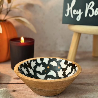 Wooden bowl with a black and white ghost pattern on the inside, styled on a tabletop with a lit candle, an orange pumpkin, and a sign in the background.