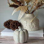 Decorative ceramic pumpkin on a wooden surface with a vase and pinecones in the background.