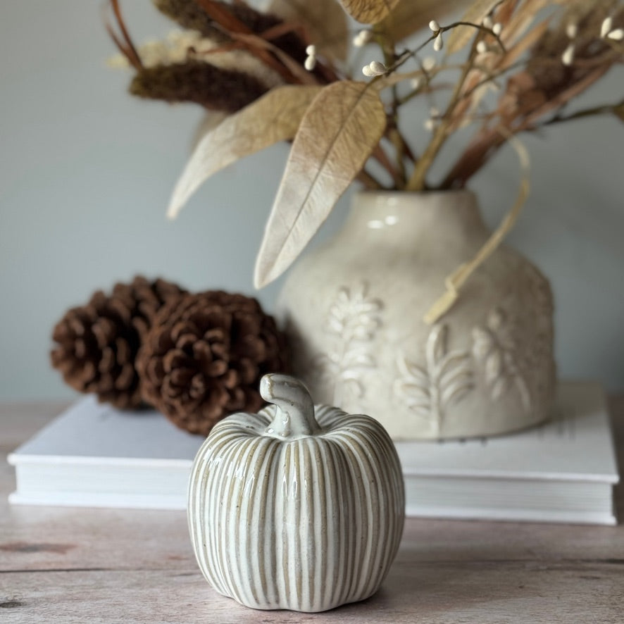 Decorative ceramic pumpkin on a wooden surface with a vase and pinecones in the background.