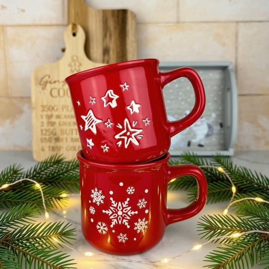 Two red ceramic mugs stacked on top of each other, decorated with white star and snowflake patterns. The mugs are styled on a kitchen counter with pine branches, fairy lights, and a wooden cutting board in the background.