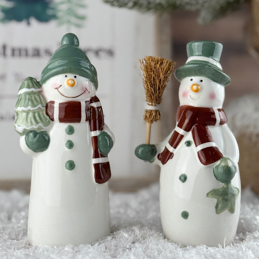 Two ceramic snowmen figurines with green hats and scarves, one holding a broom and the other a tree, on a snowy surface.