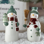 Two ceramic snowmen figurines with green hats and scarves, one holding a broom and the other a tree, on a snowy surface.