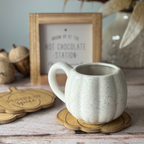 White speckled ceramic mug on a wooden coaster with 'Hot Chocolate Station' sign and decor in the background