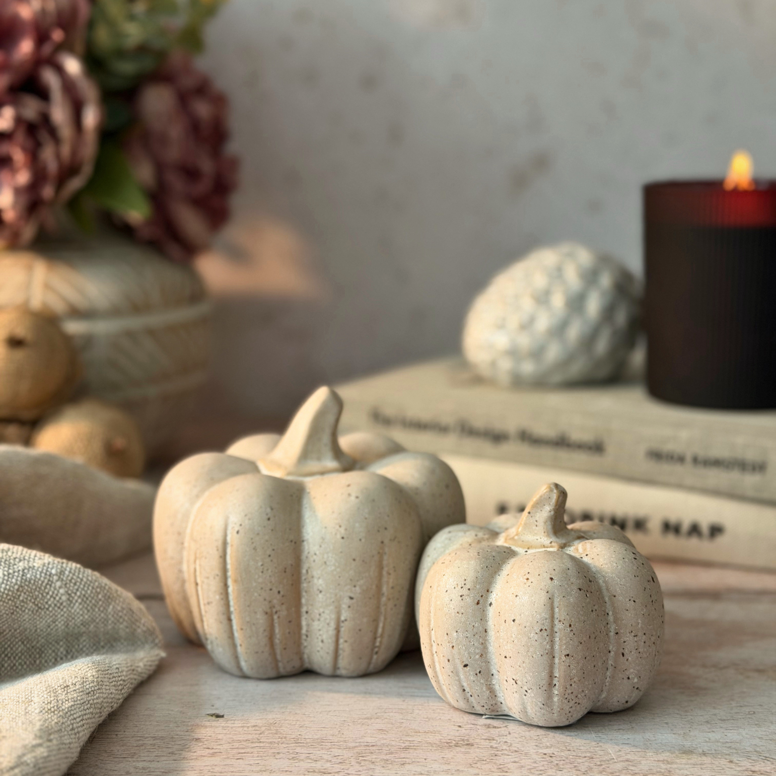 Two small decorative faux pumpkins in a speckled cream-beige ceramic finish, styled on a tabletop with a candle, books, and dried flowers in the background.