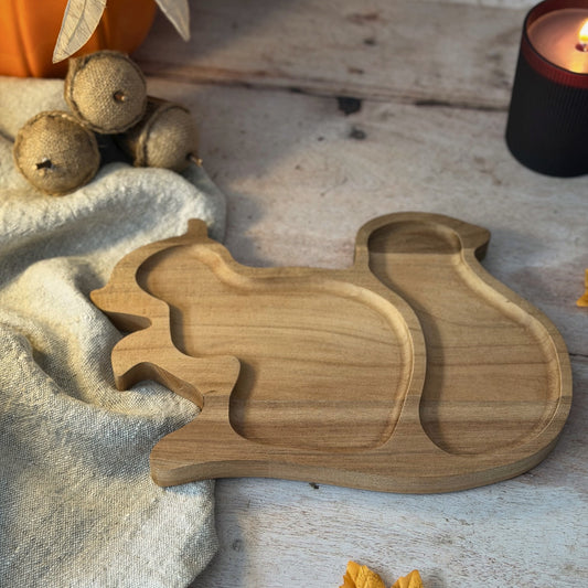 A wooden squirrel-shaped serving tray with three compartments, styled on a rustic white wooden surface with a beige cloth, acorns, a pumpkin, and a lit candle.