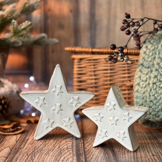 Two white ceramic stars with embossed star patterns, displayed on a rustic wooden surface with holiday decorations in the background.