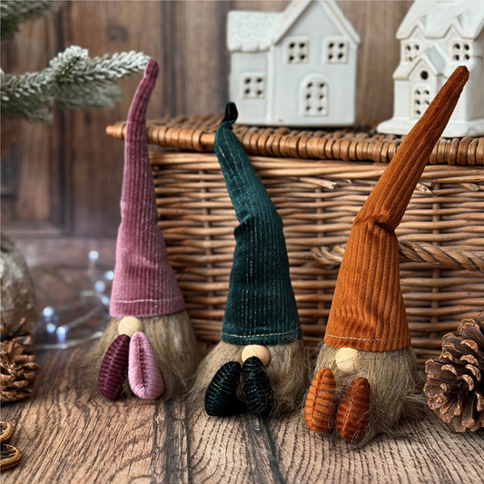 Three small corduroy gonk decorations arranged on a wooden surface in front of a wicker basket, with decorative houses, pinecones, and evergreen branches in the background.