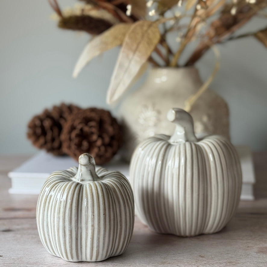 Two ceramic pumpkins on a surface with a vase and pinecones in the background.