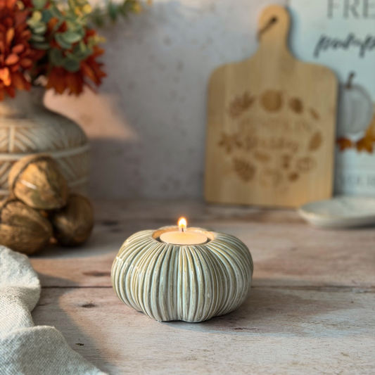 A small lit tealight candle holder shaped like a ribbed pumpkin sits on a rustic wooden table, with autumn-themed decor blurred in the background including a cutting board, pumpkins, and dried flowers.