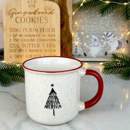 A speckled white ceramic mug with a red handle and rim, featuring a black Christmas tree graphic on the front. The mug is styled on a marble surface with festive props, including a wooden gingerbread cookie recipe sign and a framed holiday print with warm string lights.
