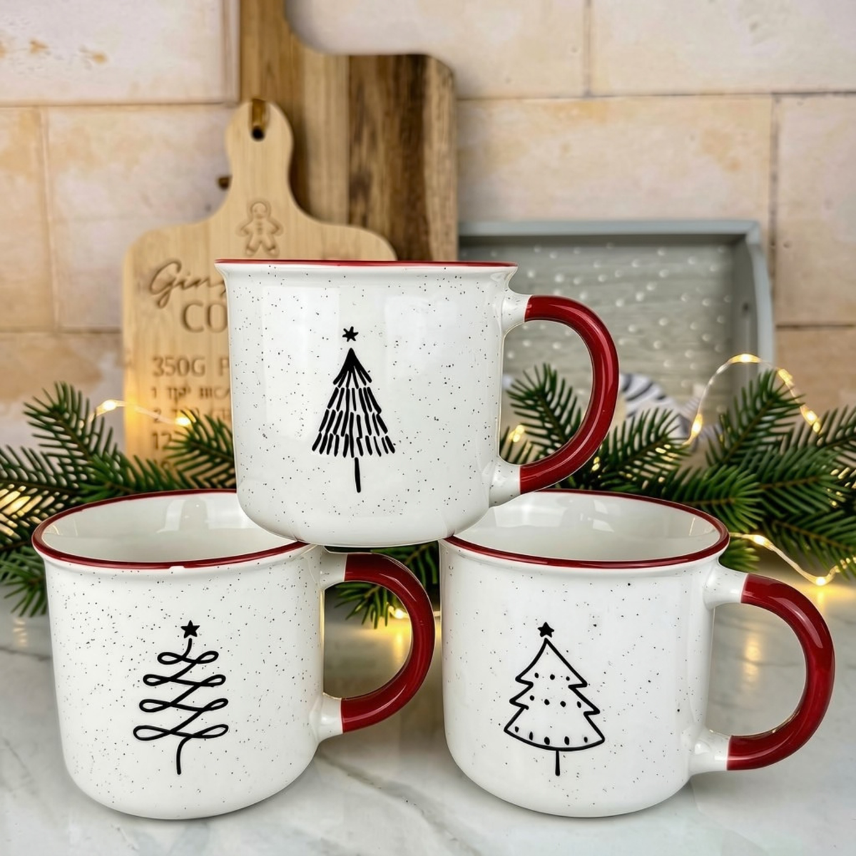Three white speckled ceramic mugs with red handles and rims, each decorated with a simple black Christmas tree design, arranged on a marble surface with holiday greenery and warm string lights in the background.