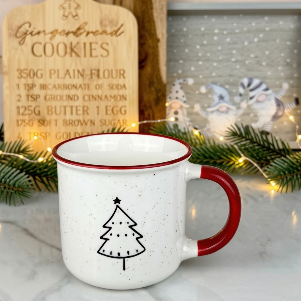 A white speckled enamel mug with a red handle and rim, featuring a black line-art Christmas tree on the front. The mug is styled on a marble surface with a wooden gingerbread cookie recipe sign, green pine branches, and warm fairy lights in the background.