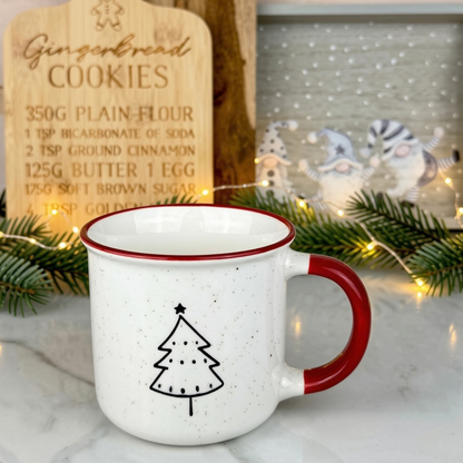 A white speckled enamel mug with a red handle and rim, featuring a black line-art Christmas tree on the front. The mug is styled on a marble surface with a wooden gingerbread cookie recipe sign, green pine branches, and warm fairy lights in the background.