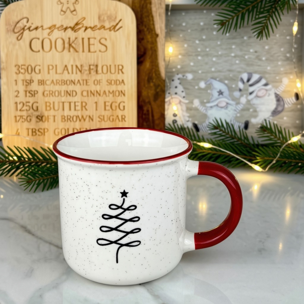 A white speckled enamel mug with a red rim and red handle, featuring a black Christmas tree design on the front. The mug is styled on a marble surface with a wooden gingerbread cookie recipe book and a festive holiday sign in the background.