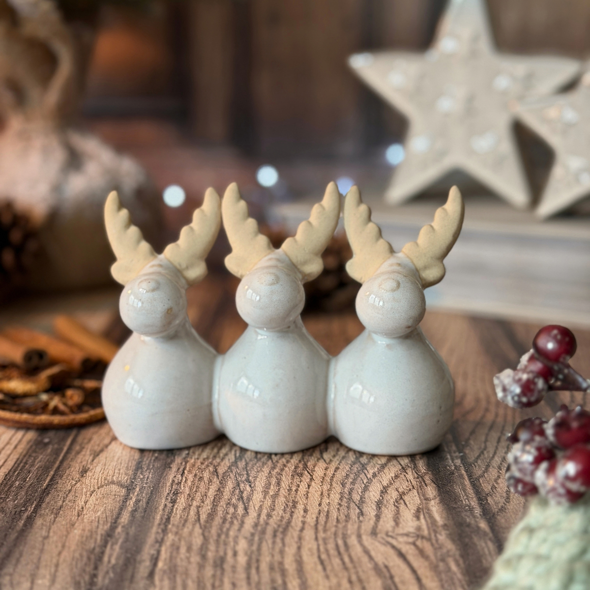 Three small ceramic reindeer figurines with cream antlers, arranged side by side on a rustic wooden surface with a warm, blurred holiday background.