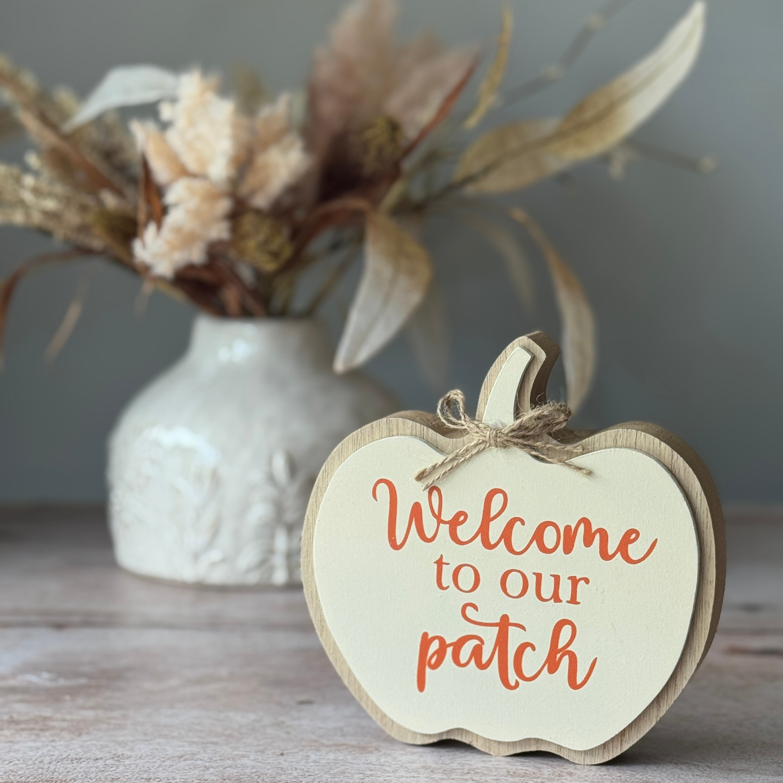 Wooden sign with 'Welcome to our patch' text on a wooden surface with a vase of flowers in the background.