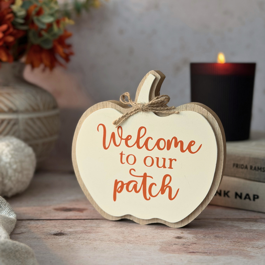 A wooden apple-shaped sign with the phrase 'Welcome to our patch' in orange script, styled on a rustic tabletop with a lit candle, books, and autumn decor in the background.