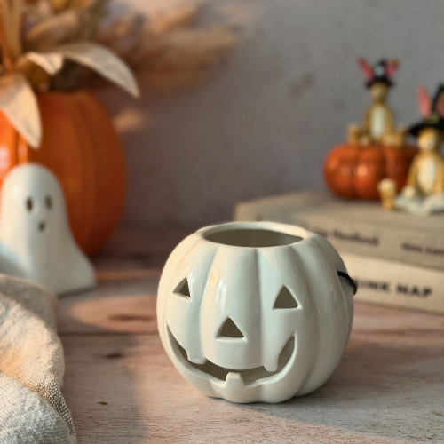 A small white ceramic jack-o'-lantern candle holder with carved facial features, styled on a wooden surface with autumn-themed props including pumpkins, a ghost figurine, and books in the background.