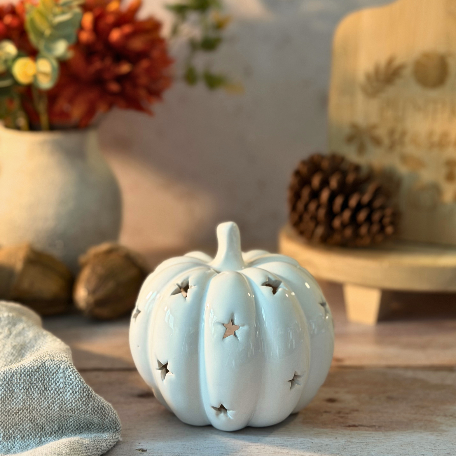 A small glossy white ceramic pumpkin ornament with star-shaped cutouts, displayed on a rustic wooden surface with autumn decor in the background.