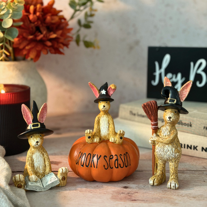 Three decorative golden rabbits wearing black witch hats are arranged on a tabletop with a small orange pumpkin that reads “Hey Boo” One rabbit sits on the pumpkin, one reads a book, and one holds a broom.