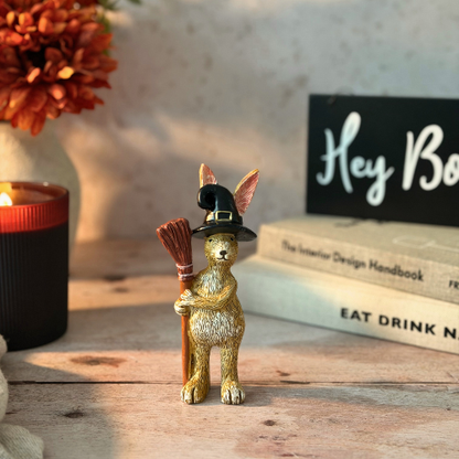 A small decorative rabbit figurine in metallic gold, wearing a black witch hat and holding a broom, displayed on a wooden surface with a candle, flowers, books, and a sign in the background.