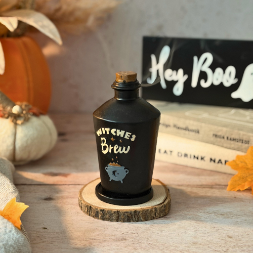 A black incense burner designed to look like a 'Witches Brew' potion bottle, with a label that includes the text 'WITCHES BREW' and an illustration of a witch's hat and a cauldron. The bottle is placed on a wooden surface with a pumpkin and incense cones nearby.