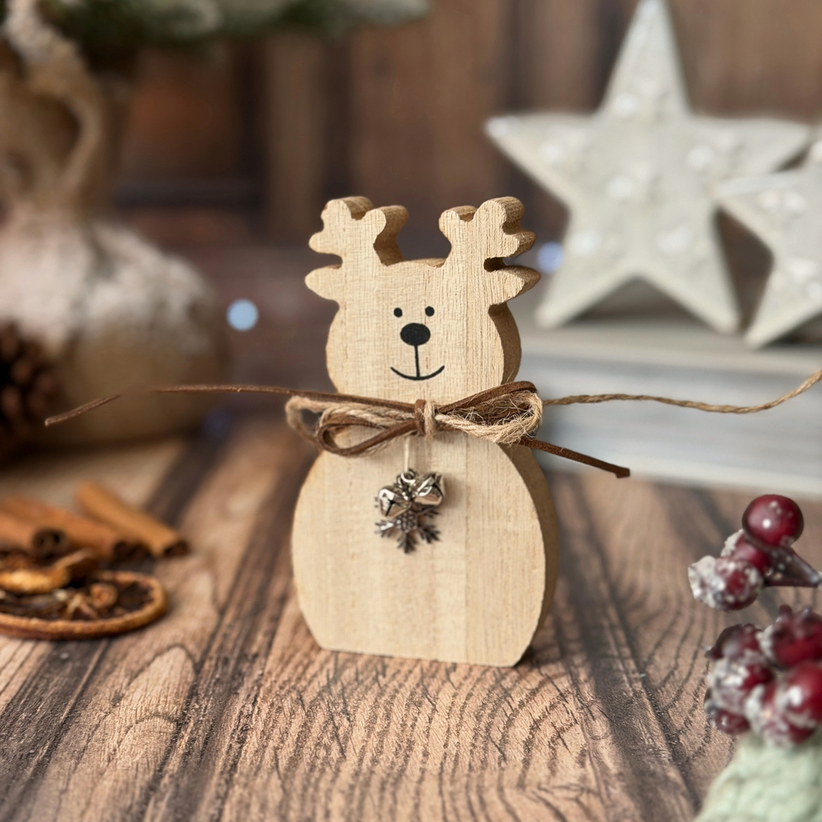 A small wooden reindeer decoration with antlers, a twine bow, and a silver bell ornament, displayed on a rustic wooden surface with holiday props in the background.