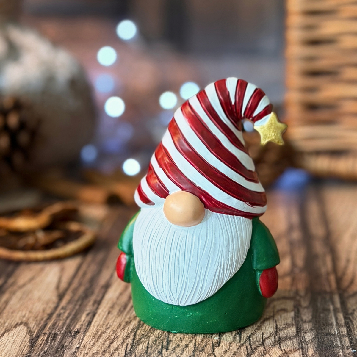 A small Christmas gonk figurine with a red and white striped hat, white beard, green outfit, and red gloves, standing on a wooden surface with a softly blurred holiday background.