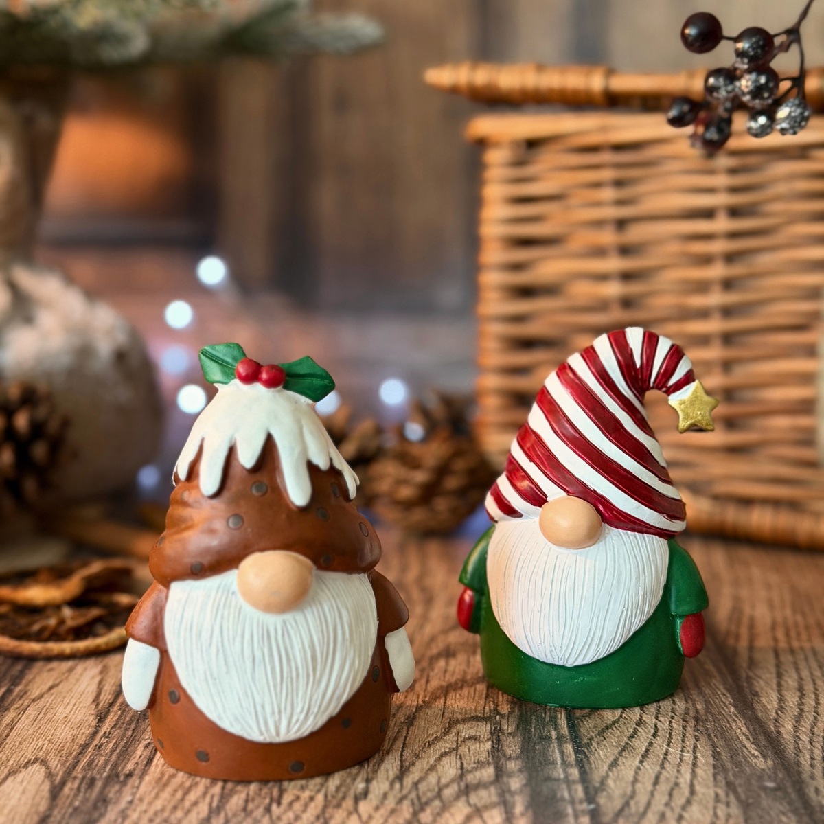 Two Christmas gonks figurines on a rustic wooden surface, one shaped like a Christmas pudding man with a white beard and dripping icing, and the other wearing a red-and-white striped hat and green outfit. Decorative props including a wicker basket, pinecones, and soft lights are blurred in the background.
