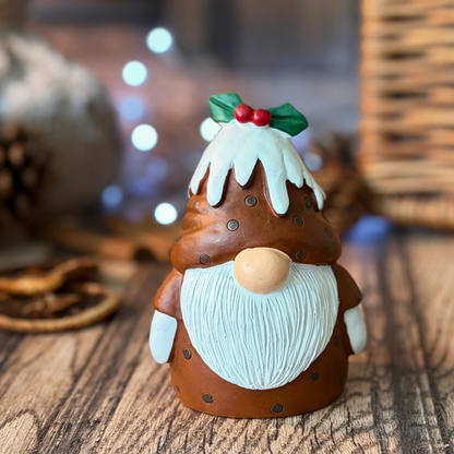 A small Christmas gonk figurine shaped like a Christmas pudding man with a white beard, brown body, and a white frosted hat decorated with green leaves and red berries, displayed on a wooden surface with a softly blurred holiday background.
